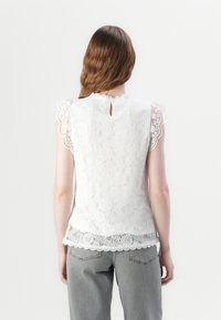 Vitt spetsblus med korta, utsvängda ärmar, scalloperade kanter och en nyckelhålbakdetalj, matchad med ljusgrå jeans.
