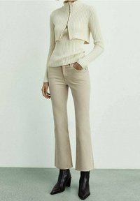 Personne portant un pull zippé côtelé beige raccourci, un pantalon évasé taille haute assorti et des bottines à talons noires, debout sur un sol vert.