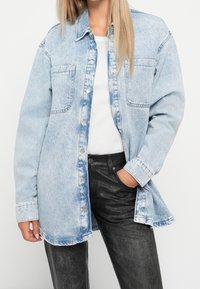 Žena v svetle modré džínové bundě s acid-wash efektem přes bílý top, s černými strukturovanými kalhotami, s jednou rukou v kapse a vlasy až po ramena.