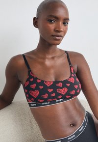 Svart bralette med rødt hjerte-mønster, justerbare stropper og hvit merket elastikk. Myk stoff med en tettsittende design, uten padding.