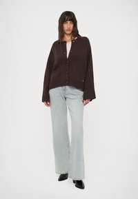 Femeie în picioare, purtând un cardigan închis la culoare cu nasturi, blugi deschiși cu picior larg și cizme negre cu vârf ascuțit pe un fundal alb.