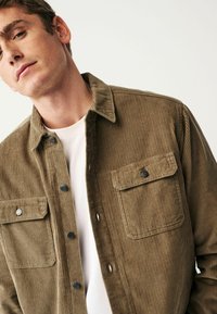 Corduroy shirt in brown, featuring a collar, button closures, two front pockets, and textured ridges throughout. Worn over a white shirt.