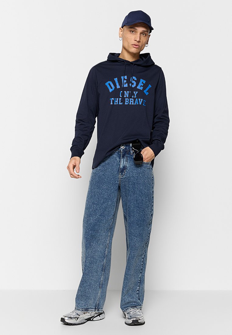 Marineblauwe hoodie met lange mouwen met blauwe grafische tekst, gecombineerd met loszittende blauwe spijkerbroeken en grijze sneakers, geaccessorieerd met een zonnebril.