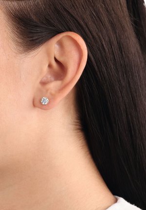 Primo piano di un orecchio femminile con capelli castano scuro lisci, che indossa un piccolo orecchino a perla di diamante tondo.