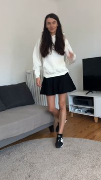 Sweatshirt branco com corte oversized, saia preta plissada e ténis pretos com detalhes brancos. Em pé num tapete claro perto de um sofá e de uma televisão.