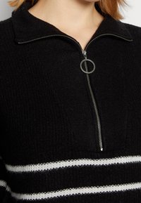 Svart stickad tröja med halvmått krage, en texturerad yta och två horisontella vita ränder över bröstet.