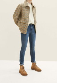 Texturovaná tvídová bunda v béžové a hnědé barvě, krémový svetr, modré úzké džíny a hnědé šněrovací boty s gumovou podrážkou.