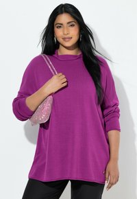 Frau mit langen schwarzen Haaren, die ein lila Langarmshirt und schwarze Hose trägt und eine pinke Handtasche mit Strasssteinen über der Schulter hält.