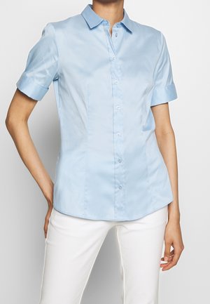 Mujer vestida con camisa de color azul claro, de manga corta, con botones y cuello, combinada con pantalones blancos, de pie con una mano en la cadera sobre un fondo liso.