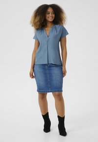 Haut bleu à manches courtes avec un col en V et un détail froncé, associé à une jupe crayon en denim bleu clair. Le mannequin porte des bottes noires à chevilles.