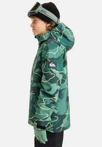 Jachetă de schi camuflaj în nuanțe de verde, cu țesătură texturată, accente galbene, izolație avansată și patch cu logo pe mânecă. Asortată cu mănuși.