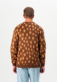 Man in een bruine fuzzy trui met oranje vlekken en lichtblauwe spijkerbroek met witte stippen, met zijn rug naar de camera gericht tegen een effen witte achtergrond.