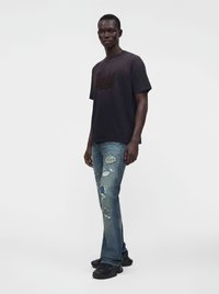 Άνδρας που φοράει μαύρο γραφικό t-shirt, φθαρμένα μπλε τζιν με patches και μαύρα αθλητικά παπούτσια, στέκεται μπροστά σε απλό ανοιχτό φόντο.