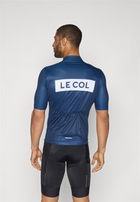 Maillot de cyclisme bleu marine à manches courtes, avec un panneau en maille respirant et un grand logo blanc "LE COL" au dos. Associé à un short noir.