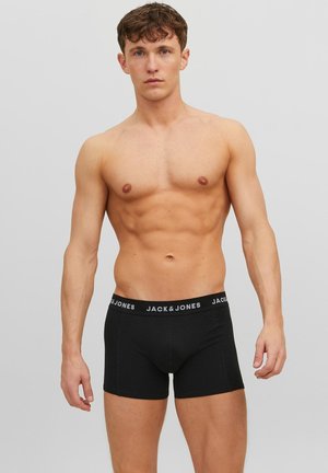 Jonge man met krullend bruin haar, gekleed in zwarte Jack & Jones boxer briefs, staand tegen een effen grijze achtergrond.