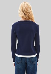 Eine blonde Person trägt ein langärmliges navy-blaues Oberteil über einem weißen Hemd und blauen Jeans und steht mit dem Rücken zu einem schlichten grauen Hintergrund.
