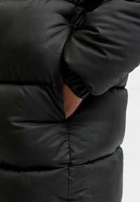 Melnā pūķa jaka ar kvadrātveida šuvēm, gludu tekstūru un elastīgām piedurknēm; ietver sānu kabatu.