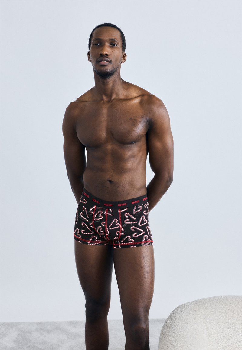 Μαύρα αντρικά μποξέρ με σχέδια γλυφιτζούρια σε κόκκινο και λευκό, με μαύρη ελαστική μέση με λογότυπο. Το μοντέλο στέκεται μπροστά σε φωτεινό φόντο.