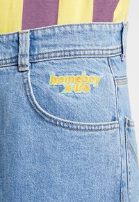 Helle blaue Denim-Jeans mit orange-gelbem gesticktem "homeboy x-tra"-Label und einem geschwungenen Design der Gesäßtasche. Abgeschlossen mit sichtbaren Nähten.