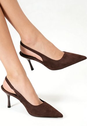 Pieds portant des escarpins slingback en daim marron foncé avec talons aiguilles plats et bouts pointus sur fond blanc.
