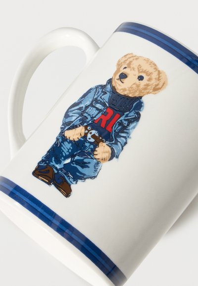 Λευκή κεραμική κούπα με μπλε λωρίδες στην άκρη, που απεικονίζει μια αρκουδίτσα ντυμένη με μπλε μπουφάν, τζιν και κόκκινο πουλόβερ "RL", κρατώντας ένα παιχνίδι.