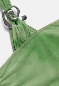 Gros plan d’un vêtement en tissu maille verte avec un détail de fermoir en métal argenté torsadé sur une bretelle sur fond blanc.