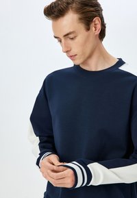 Mörkblå sweatshirt med kontrasterande vita ärmar, ribbade mudd med marinblå och vita ränder, samt rund halsringning. Slät tygkvalitet.