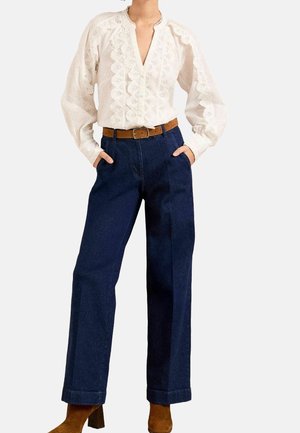 Persoon draagt een witte blouse met kantpatroon, ingestopt in een hooggetailleerde donkerblauwe wijde broek, met een bruine riem en bruine laarzen.