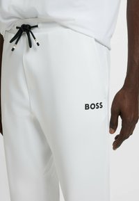 Witte sportbroek van glad materiaal, voorzien van een tailleband met trekkoord en een gedrukt zwart "BOSS" logo op de linker bovenbeen.