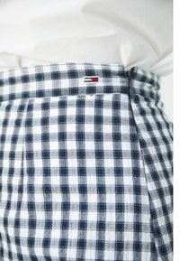 Gros plan sur une taille portant un pantalon à carreaux bleu et blanc avec un petit logo rouge et blanc sur la ceinture et une chemise blanche rentrée.