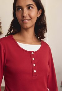 Red long-sleeve top with a round neckline and five white buttons down the front. Features a soft, smooth texture and layered white shirt underneath.