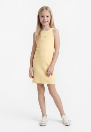 Ragazza giovane in piedi, indossa un vestito giallo senza maniche con una piccola farfalla rosa sul petto e scarpe da ginnastica bianche.