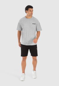 Grå bomulds T-shirt med korte ærmer, der har et sort logo "SMILODOOK" på venstre bryst. Bæres med sorte shorts og hvide sneakers.