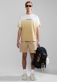 Lys gradient t-shirt, der transitions fra hvid til beige, parret med beige shorts, hvide sokker og beige sneakers. Sort rygsæk med logo.