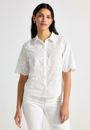Hvid kortærmet bluse med blomsterbroderi og hulmønsterdetaljer. Knappelukning med en bølget kant forneden og ved kraven.