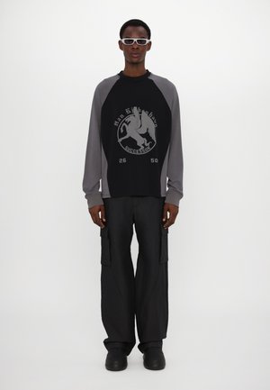 Czarna i szara koszulka raglanowa z długim rękawem z graficznym nadrukiem, zestawiona z szerokimi czarnymi spodniami cargo i czarnymi butami. Białe prostokątne okulary przeciwsłoneczne.