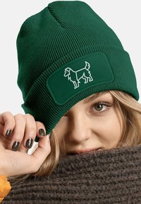 Green knit beanie with ribbed texture and a rectangular patch featuring a white outline of a dog. The fabric appears soft and flexible.