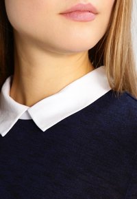 Mörkblå tröja med vit krage. Har en texturerad kvalitet och en avslappnad passform, vilket framhäver kontrasten mellan kragen och kroppen.