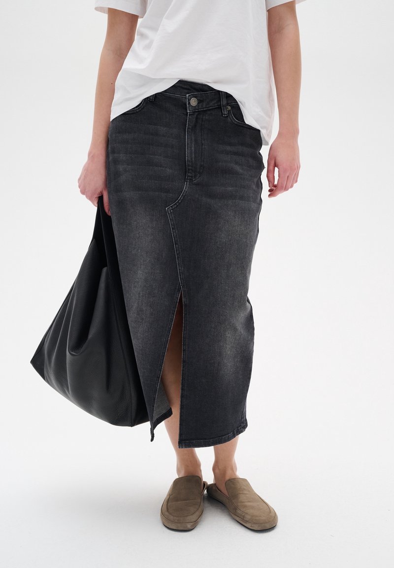 Black denim skirt with a front slit, high waist, and contrasting stitching, paired with a loose white t-shirt and brown slip-on shoes.