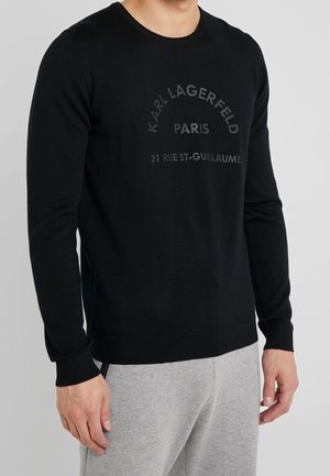 Schwarzer Strickpullover mit rundem Ausschnitt, langen Ärmeln und einem Logodruck in Dunkelgrau. Gerippte Bündchen und Saum. Kombiniert mit grauen Hosen.
