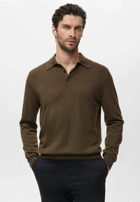 Hombre con cabello oscuro que lleva un suéter de polo de manga larga marrón y pantalones oscuros, de pie con las manos en los bolsillos frente a un fondo liso.
