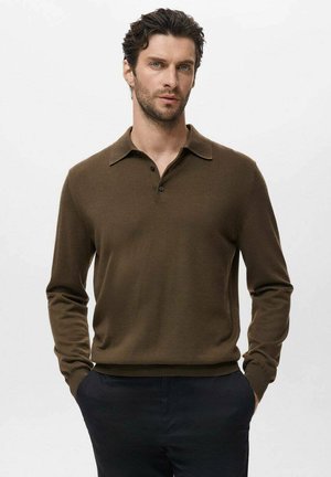 Man met donker haar, gekleed in een bruine polo met lange mouwen en donkere broek, staand met handen in de zakken tegen een effen achtergrond.