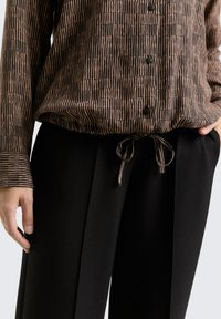 Braun und schwarz gestreifte Bluse mit langen Ärmeln, Knopfverschluss und einem Band in der Taille, kombiniert mit hoch taillierten schwarzen Hosen.