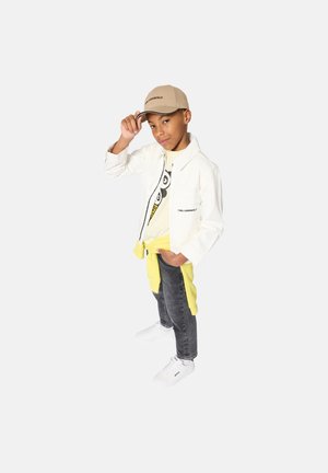 White denim jacket, yellow sweatshirt tied at the waist, gray jeans, and white sneakers. Tan cap with logo on front; child posed playfully.