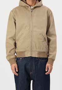 Tan zip-up hoodie with a hood, front pockets, and ribbed cuffs. Features a smooth material and simple design; paired with dark denim jeans.