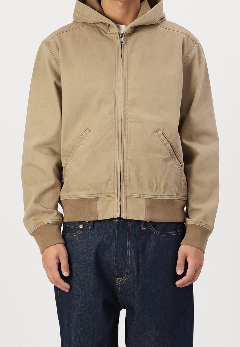 Tan zip-up hoodie with a hood, front pockets, and ribbed cuffs. Features a smooth material and simple design; paired with dark denim jeans.