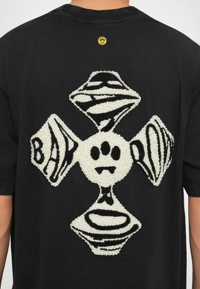 Svart t-skjorte med en teksturert design på baksiden, med hvite og svarte perledetaljer som danner et kors og abstrakte former.