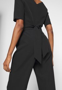 Combinaison noire à manches courtes, décolleté en V et détail nœud au dos. Caractérisée par une texture lisse et une coupe structurée.