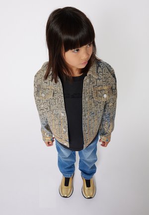 Giubbotto di jeans con una texture a motivo di blu chiaro e oro, camicia nera, jeans blu chiaro e scarpe slip-on metalliche dorate.