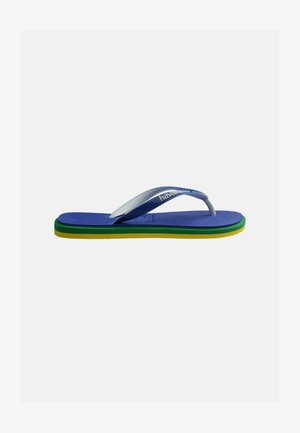Beine einer Person, die einen rosa-weiß gestreiften Rock und blaue Flip-Flops trägt, stehen auf einem schwarz-weißen Mosaikpflaster in der Nähe von roten Stühlen und einem gelb gestreiften Sonnenschirm.